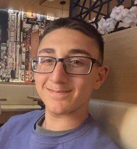 Smiling young man with short hair and rectangular glasses, wearing a purple shirt in a cozy cafe setting behind him a wall mural and seating.