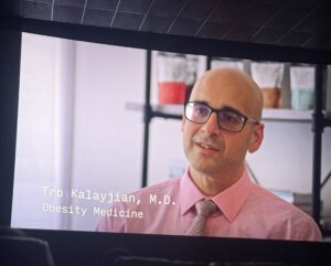Portrait of Dr. Tro Kalayjian wearing a pink shirt and glasses, speaking in a presentation screen (Obesity Medicine).