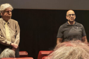 Two men sit on a stage in front of a dark backdrop; audience members are visible in the foreground.