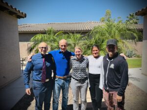 Five adults pose together outdoors in a sunny backyard with palm plants and a tiled roof house in the background, smiling for a group photo.