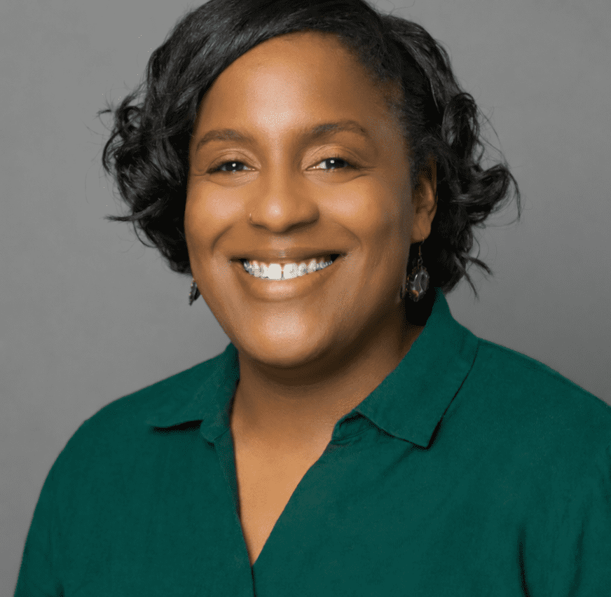 Smiling African American woman with short curly black hair, wearing a green blouse and dangling earrings against a gray backdrop.