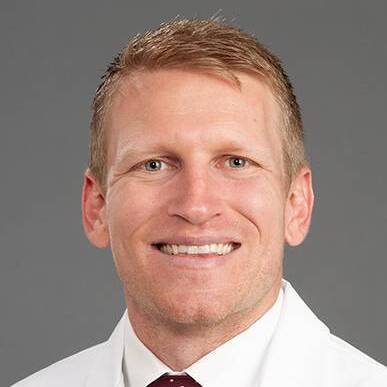 Smiling man with short blond hair in a white dress shirt and burgundy tie against a gray background (professional headshot).