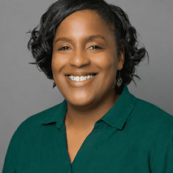 Smiling African American woman with short curly black hair, wearing a green blouse and dangling earrings against a gray backdrop.
