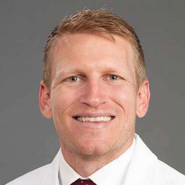 Smiling man with short blond hair in a white dress shirt and burgundy tie against a gray background (professional headshot).